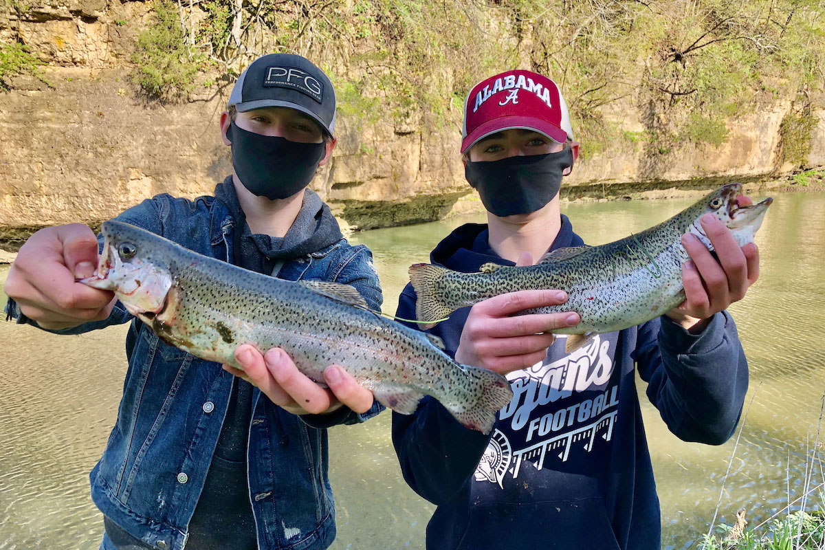 Two young men stand near the shoreline of a waterway and present their successful catch of the day speckled green and silver trout. In the background is the waterway against a rock outcropping.