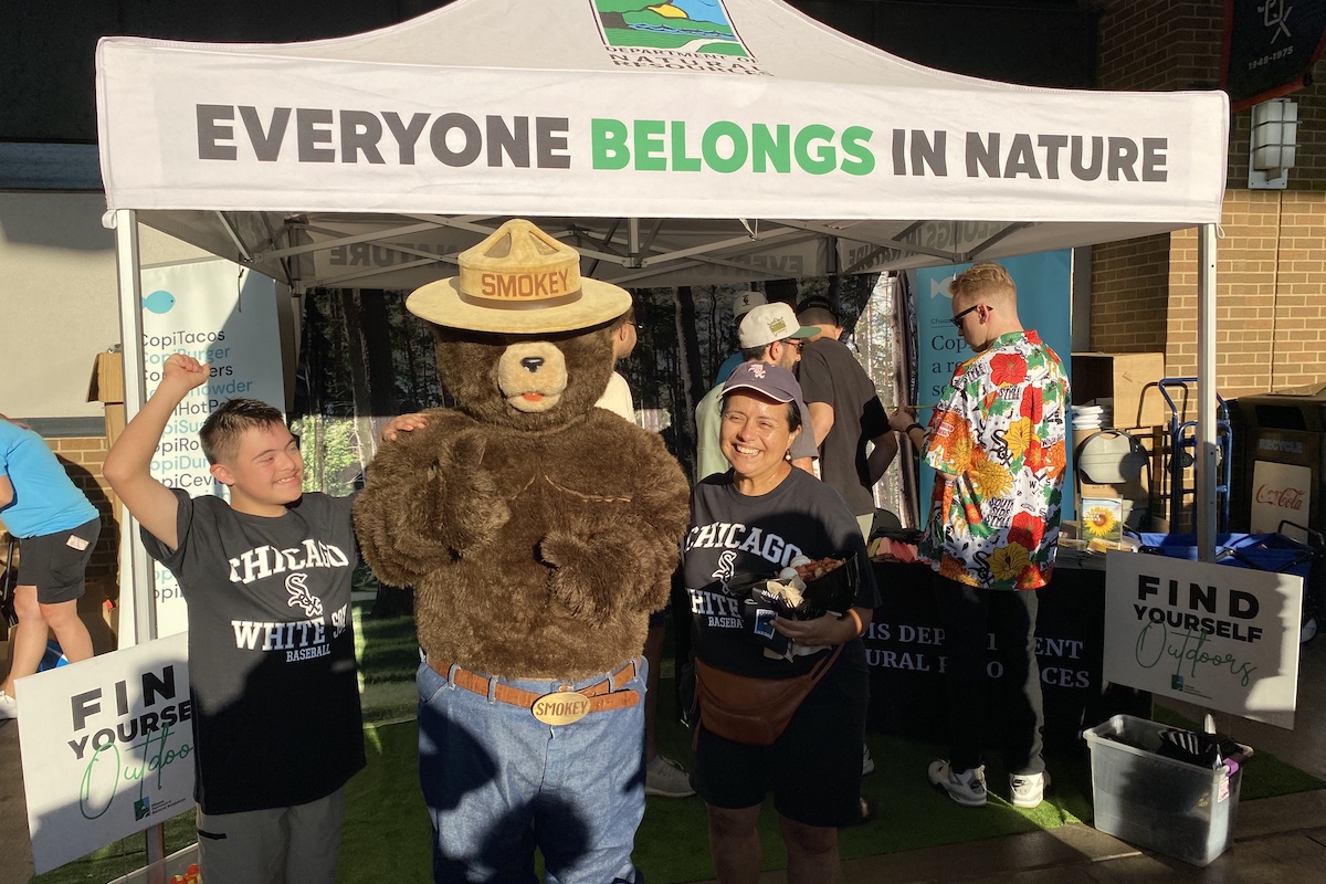 A mother and son pose next to the smokey the bear mascot while visiting the Illinois Department of Natural Resources Everyone Belongs in Nature tent. In the background are a few other folks talking to the staff and looking at the displays.