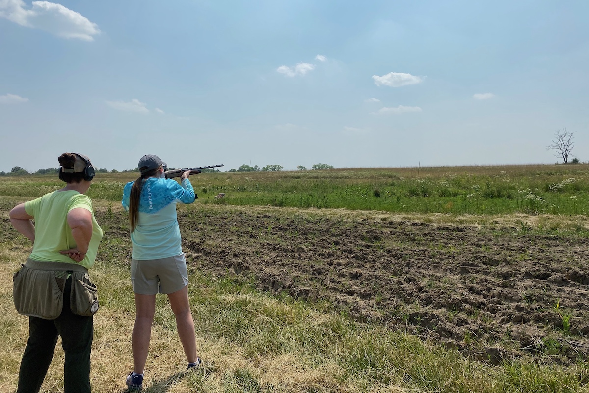 Two people stand to the left in the foreground. One person holds a shotgun up to her shoulder and aims at the bright blue sky. The second person stands behind the individual aiming the shotgun. In the background is a grassland.