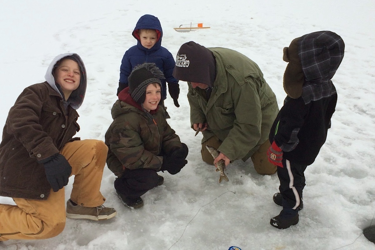 A group of children and an adult pose together on top of an ice covered lake. The adult bends over and works to remove a hook from a fish caught while ice fishing.