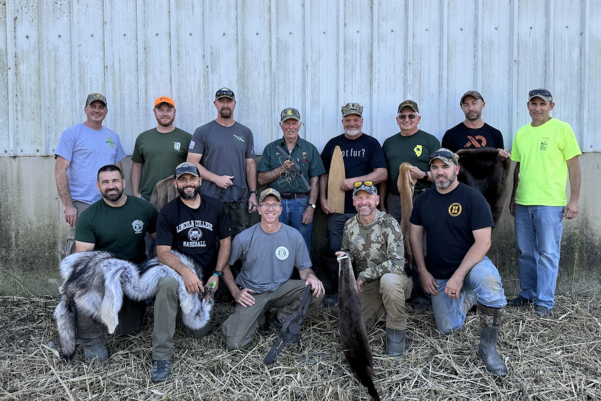 A group of men pose together against a building covered in silver metal siding against a concrete foundation.