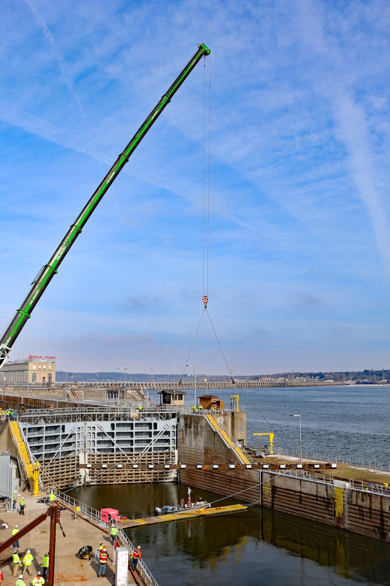 A view of a lock and dam on the Mississippi river. A crane lifts a large steel bar about to be installed in the lock approach channel. In the background is river.
