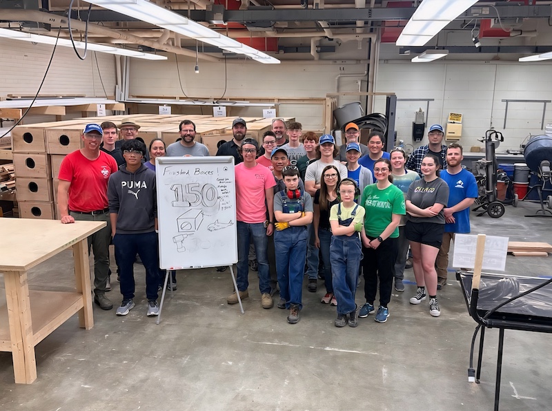 In a large room, a group of people stand together near a white board sign that says, "150 finished boxes." Behind the group of people is a large stack of wooden boxes.