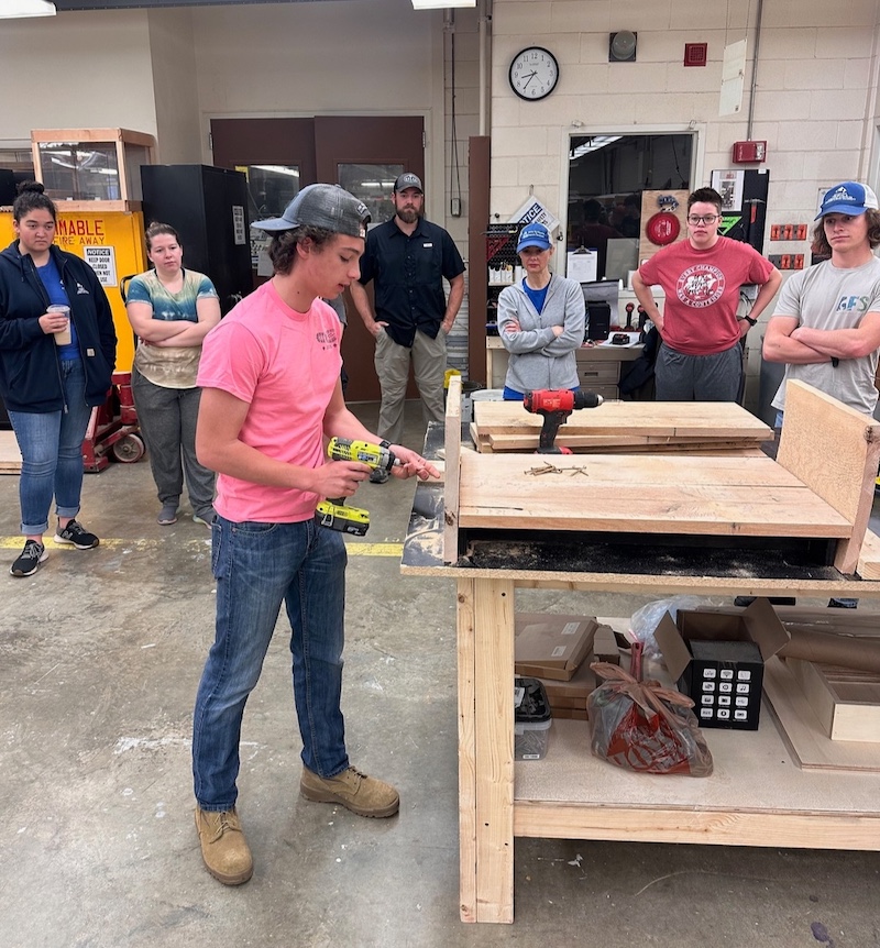 In a room with wood working tools, a group of people stand and watch as one young man conducts a demonstration of building a wooden box. The individual is holding a battery operated drill and is preparing to secure a side of the box of the wooden base. 