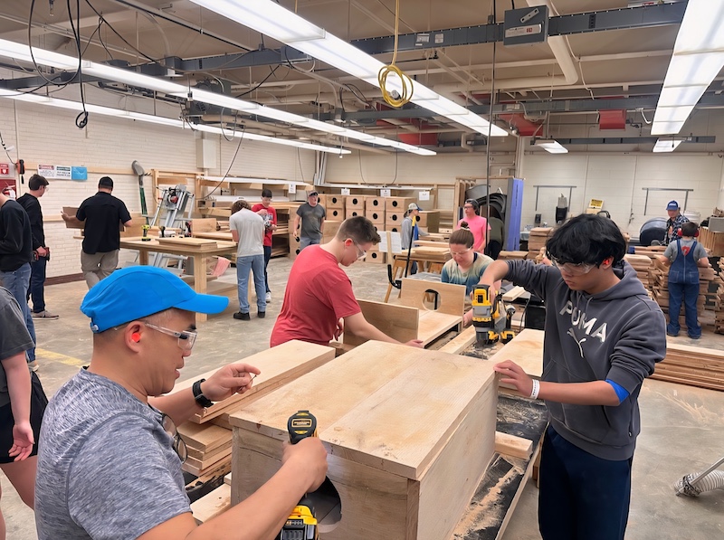Many people work together to build wooden boxes in a large room filled with tables and wood working tools.