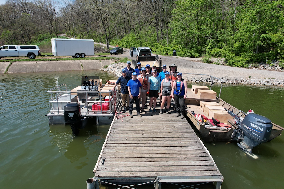 A group of people stand together on a wooden dock over the shoreline of a freshwater lake. On either side of the dock is a metal boat filled with wooden boxes. In the background is a woodland with a road winding through down to the dock.