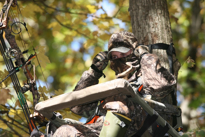 A hunter in camouflage gear sits in a tree stand attached to a tree trunk. Her compound bow is next to her. In the background is a leafy autumn canopy.
