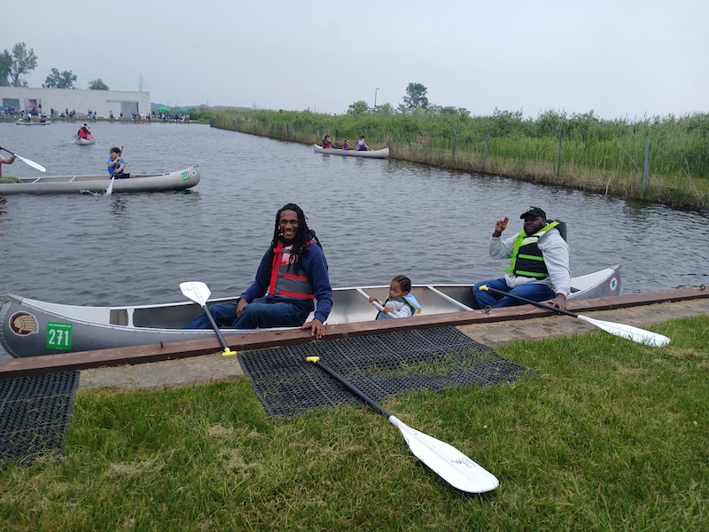 Two men and a child sit in a canoe along the side of a canal about to push off and enjoy a paddle around a pond. In the background are more people paddling around in canoes. People are fishing from the shoreline way off in the distance.