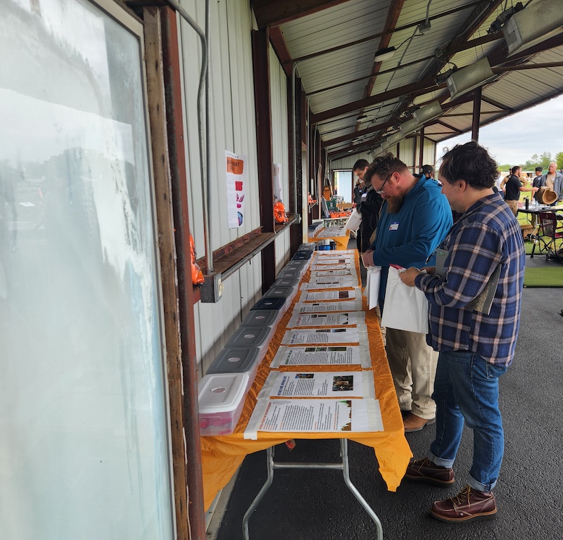A group of people visit a long row of tables in a metal sided building  open on one side. The group of people are reading documents placed on the top of the tables covered in an orange tablecloth. In the background are more people conversing with each other next to tables..