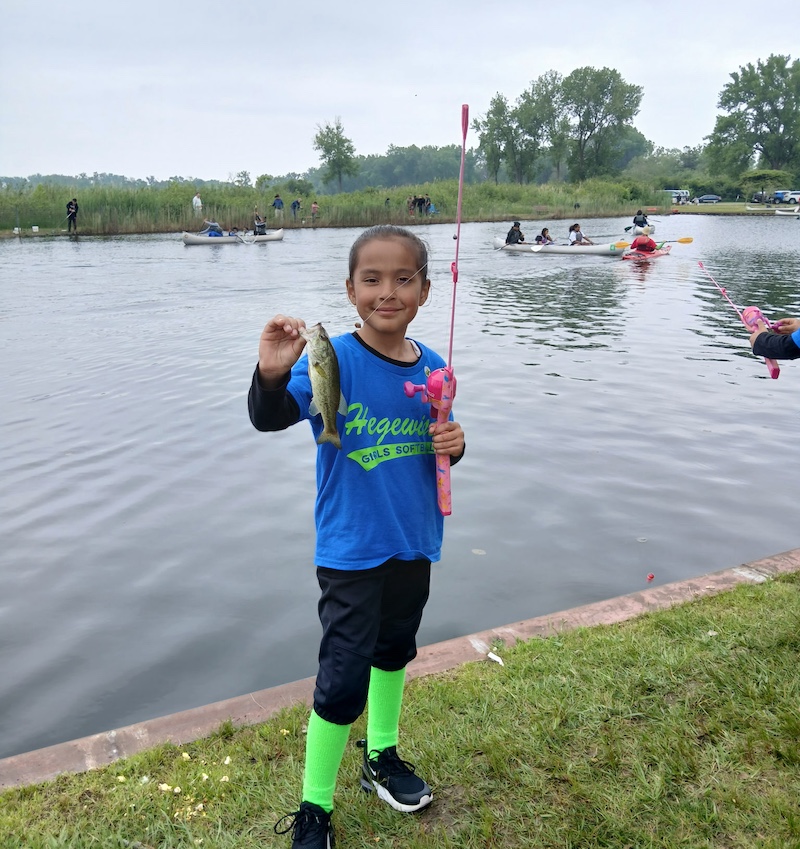 A young girl proudly holds up a silver and dark green fish on a hook attached to a fishing line connected to a fishing rod and reel in her other hand. In the background is a pond with many people and children fishing from the shoreline and some people in canoes paddling around in the middle of the pond. 