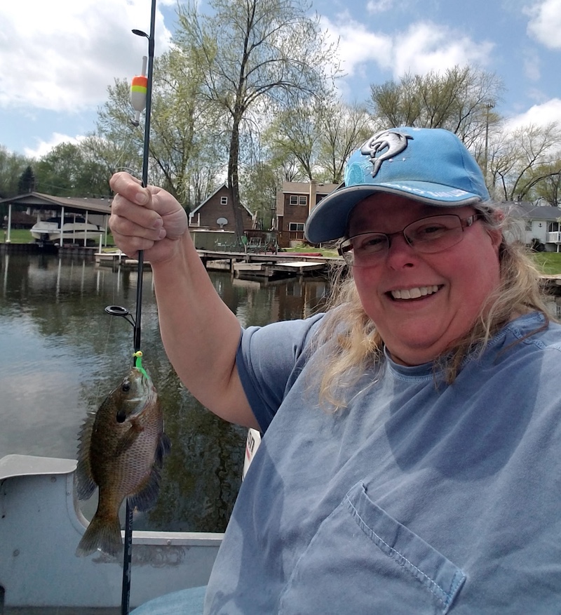 A woman in a boat on a lake poses while holding up her successfully caught small fish on a hook hanging from a fishing line attached to a rod and reel. In the background along the edge of the lake are docks connected to a line of houses.