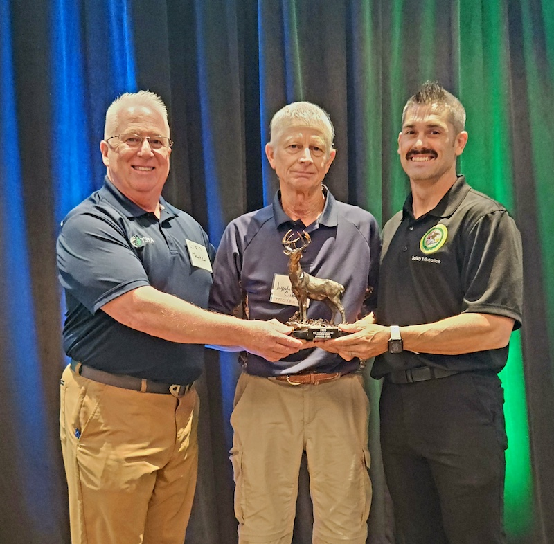 Three men pose together as the center man accepts an award. The award is a small bronze deer with antlers attached to a plaque.