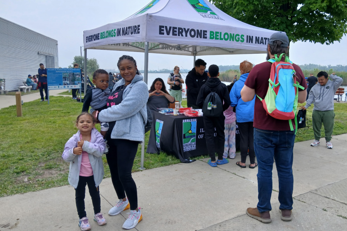 A group of people stand in front of a tent with the Illinois department of Natural Resources "Everyone Belongs in Nature logo on the canopy of the tent. A mother holds a little boy and next to her stands her little girl. A woman sitting under the tent bring hers hands together in the shape of a heart. In the background people gear up to fish at a lake.