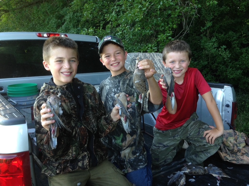 Three young boys sit on the bed of a pick up truck and pose while holding up successfully harvested mourning doves. All three boys wear camouflage gear. In the background is the silver truck parked along the edge of a woodland.