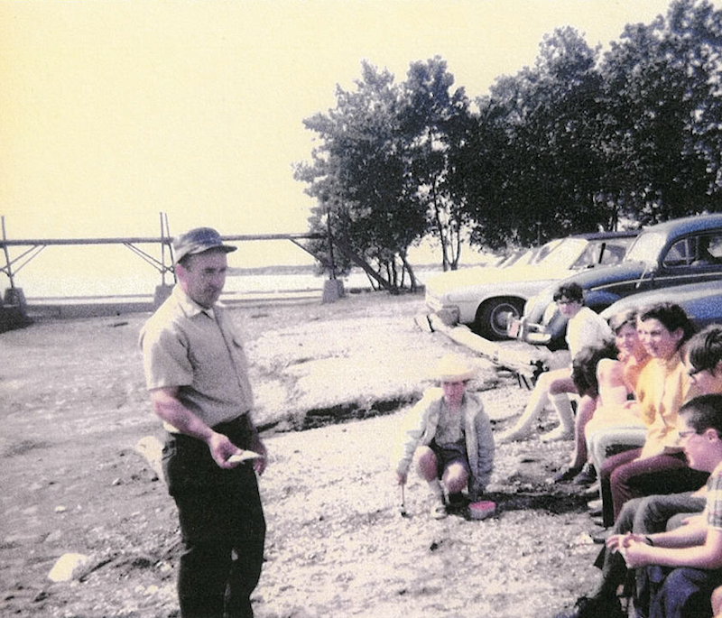 A historical photo of a fisheries biologist holding a fish and talking to school children on the shoreline of a lake. In the background are cars in a parking lot and trees.