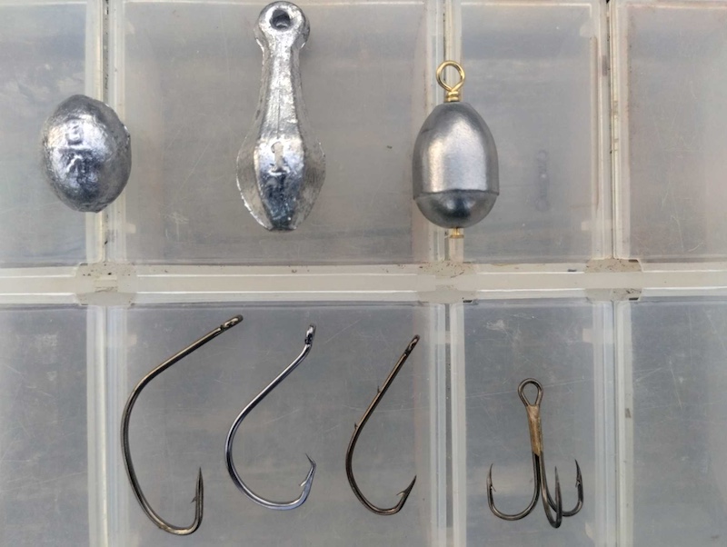 Different types of fishing hooks and silver weights used for catching catfish are lined up on top of a clear plastic tackle box.