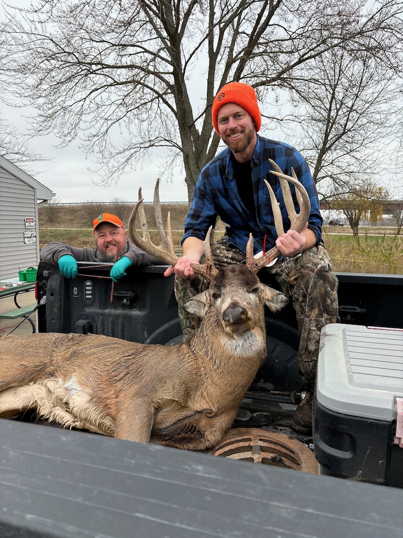 A hunter stands in the bed of a pick up truck and poses with a successfully harvested adult male white-tailed deer with large antlers. Another man leans on the side of the bed of the pick up truck and celebrates with the hunter. In the background is large tree in front of a highway.