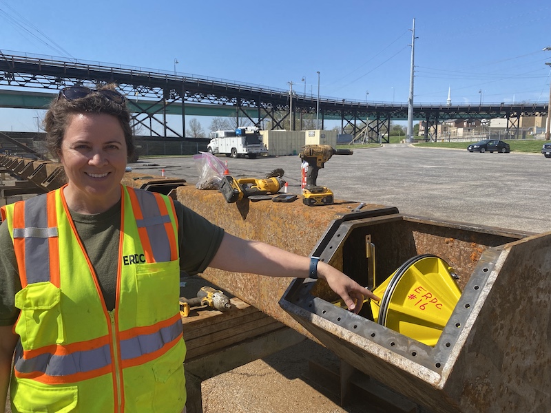 A woman in a neon yellow construction vest points to a yellow speaker inside a steel bar. In the background is a parking lot.