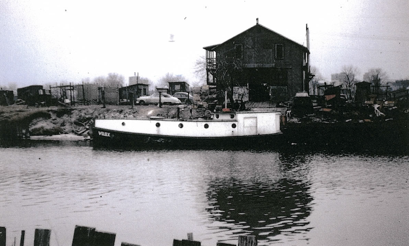 A black and white historical photo of a commercial fishing boat along the shoreline of Waukegan Harbor. A large building is in the background.