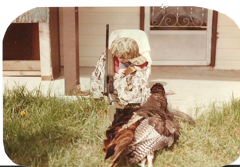 A historic photo of a young baby in a chair on a front porch surrounded by hunting gear. In the foreground is a successfully harvested wild turkey.