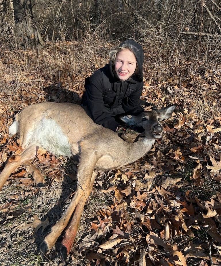 A young hunter kneels on the forest floor covered by leaf litter and poses with her successful harvested white-tailed deer. In the background is woodland.