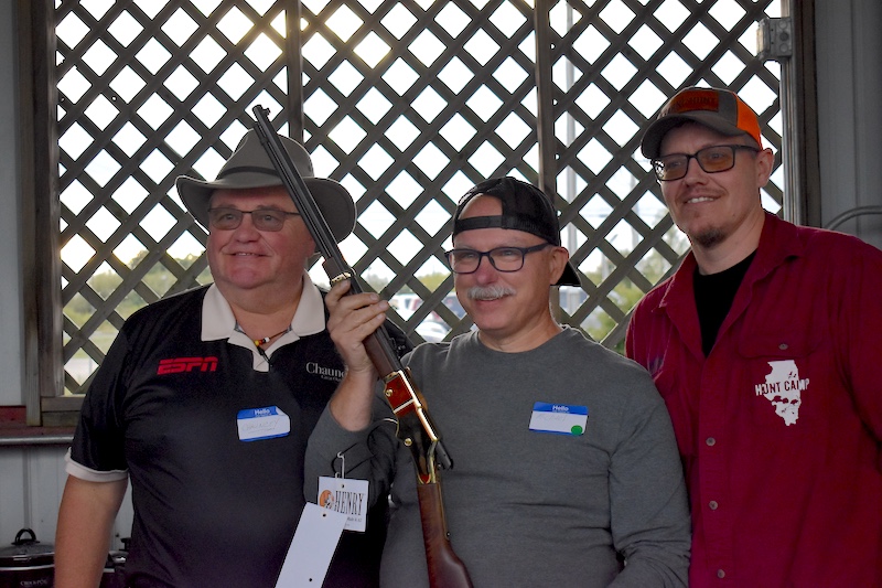 Three men standing together. The man in the middle is holding a shotgun upright and pointed towards the ceiling. In the background is a window covered in lattice.