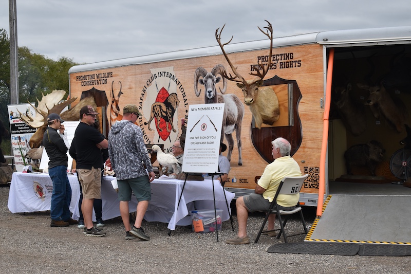Outdoors under an overcast sky, a group of people visit a display of taxidermy resting on a long table  with a white tablecloth. In the background is a walk-in trailer where individuals can see more displays.