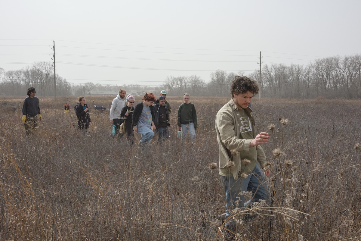 A group of people walk through an early spring grassland. Far in the background is a building surrounded by trees along the horizon.