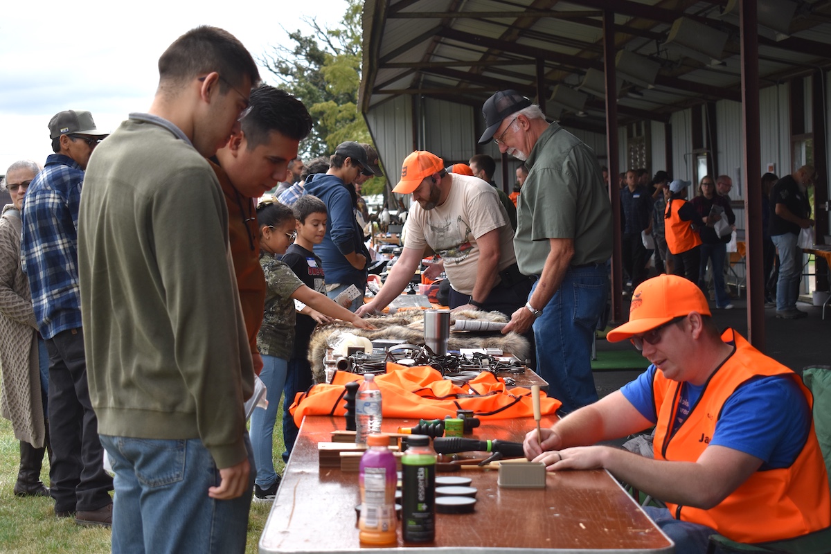 A group of adults and children visit a row of tables with an assortment of hunting gear displayed on top. The children feel and explore the different animal pelts. In the background a metal roof extends over the tables and people mill around under the shade of the roof.