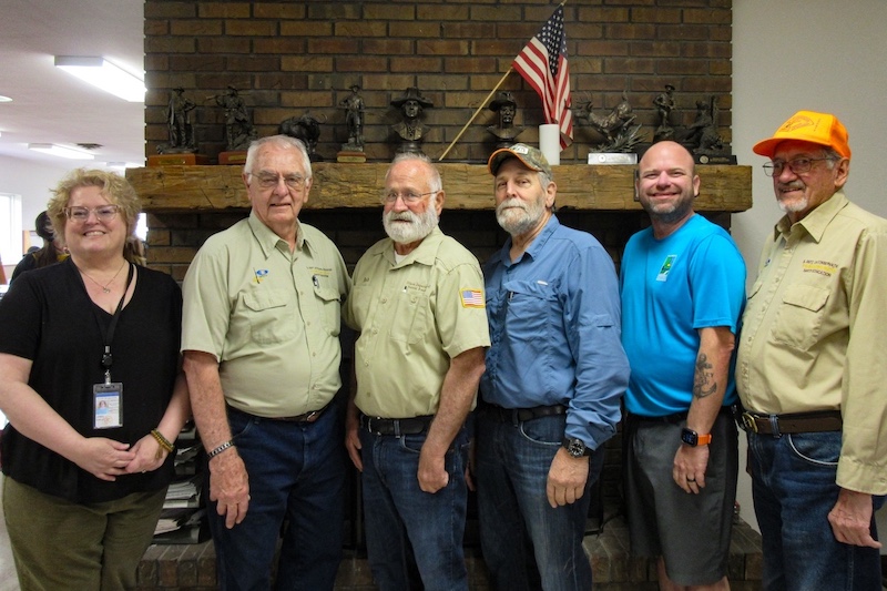 A group of men and one woman pose together in front of a brick fireplace. A rough hewn wooden beam serves as the mantle with many small bronze sculptures attached to plaques commemorate awards received rest on top.