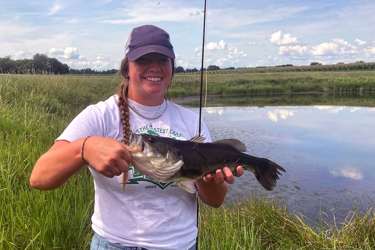 A young woman stands near a pond during a summer day under a partly cloudy blue sky and poses with her large fish just caught on a fishing line. Her fishing rod rests by her side. In the background is an agricultural field against a horizon line of trees.