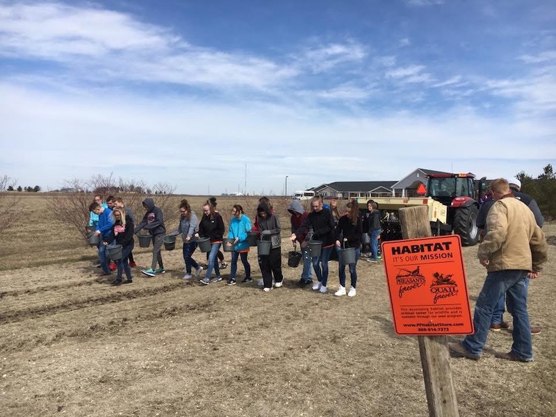 A group of people hold buckets and scatter prairie seeds by hand while walking over a tilled fallow agricultural field. In the background is a tractor with a planter attached on the back. In the distance is a large building against a bright blue partly cloudy sky.