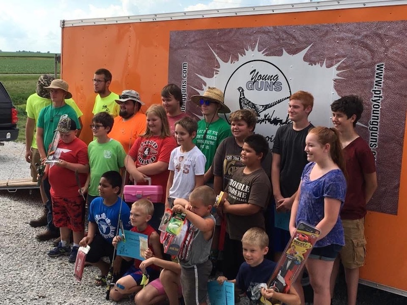 A group of youngsters stand in front of an orange trailer. In the background are green agricultural fields.