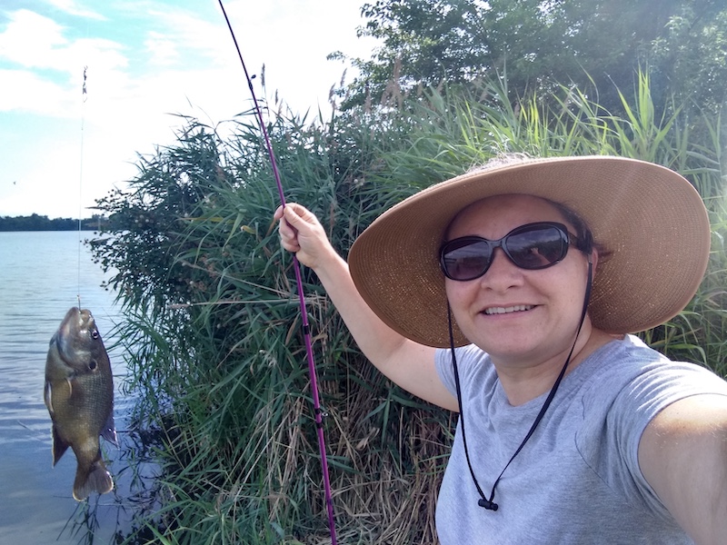 A woman poses with her successfully caught fish dangling from a hook on a line attached to a fishing rod and reel. In the background are tall grasses along the edge of a lake.