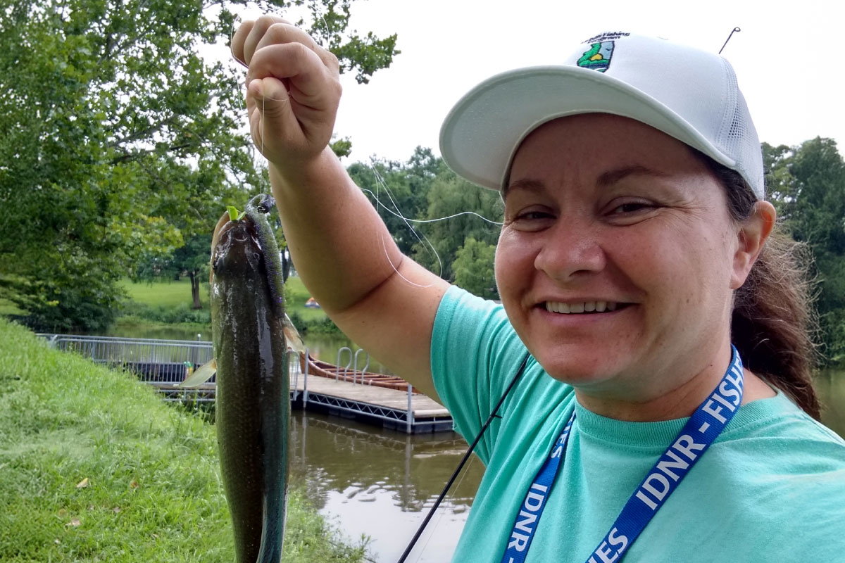 A close up of an angler holding up a fish caught on a fishing line with a lure in her right while she poses for the camera. In the background is a dock on a lake. Green trees are in the background.