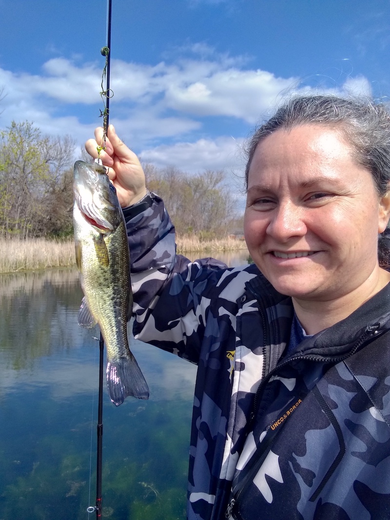 A woman holds up a fishing rod posing with her just caught green and black largemouth bass. In the background is the grassy shoreline of a pond in front of a woodland.