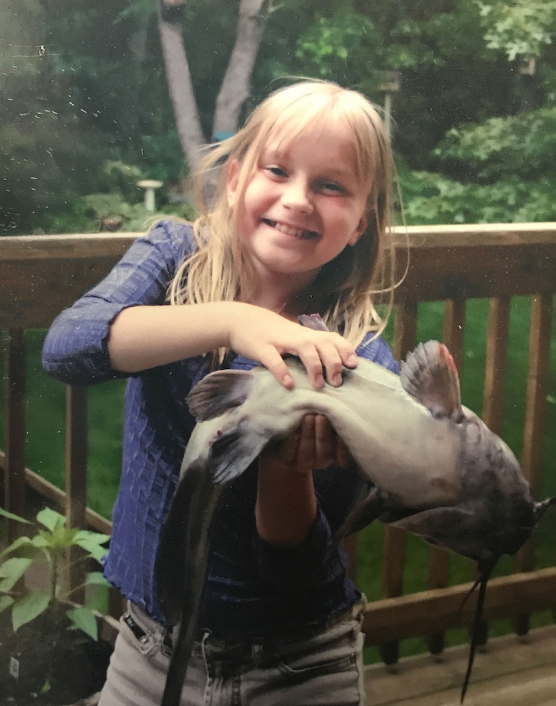 A young girl holds up her prize caught catfish. In the background is a wooden railing on a deck in front of a shady backyard along a woodland.