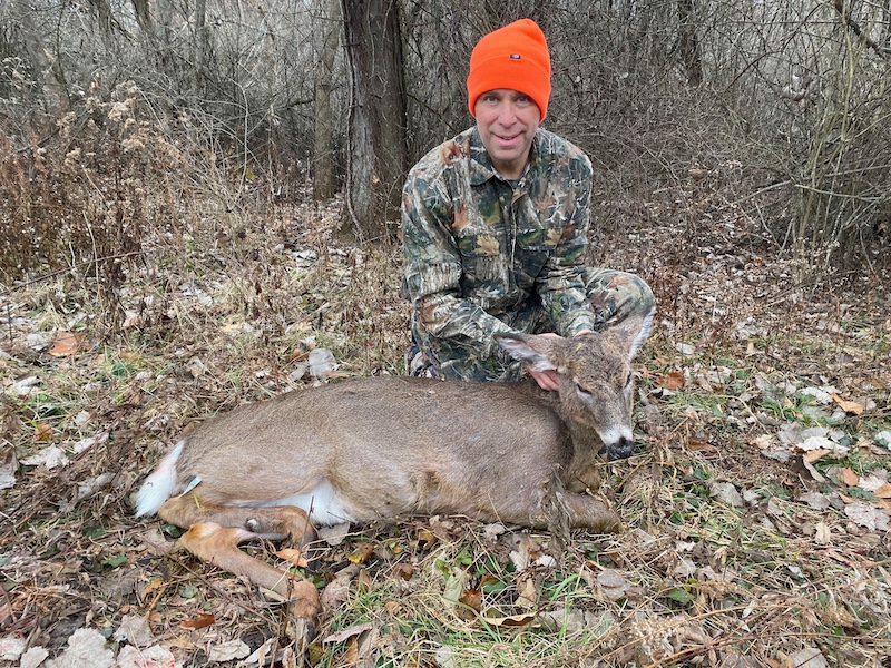 A hunter wearing camouflage gear and kneeling next to his successfully harvested white-tailed deer. In the background is a brushy woodland.