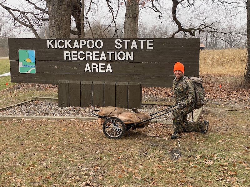 A hunter in camouflage gear pauses to kneel and pose next to a brown wooden park sign while carting out his successfully harvested white-tailed deer. In the background is an agricultural field planted to corn and left for wildlife in winter.