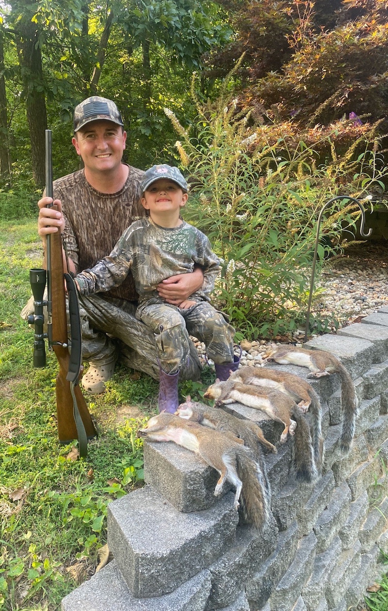 A father and daughter are dressed in camouflage gear and pose next to their successfully harvested gray squirrels resting on a concrete retaining wall. The father holds on to a rifle and in the background is a woodland.