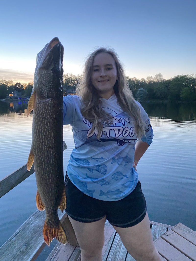 A woman standing on a wooden dock on a lake holds up a large green and white speckled fish at sunset. In the background is the shoreline of the lake with houses and trees interspersed.