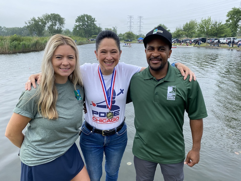 Two women and one man pose together in front of a lake at a fishing festival. People in the background are paddling around the lake in canoes or standing at the shoreline fishing.