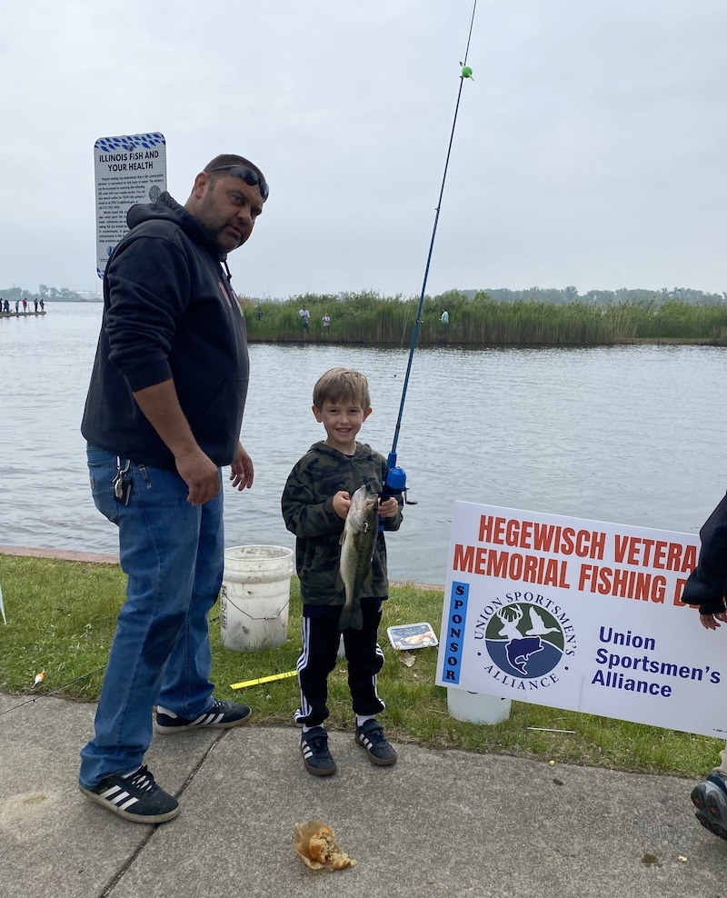 A young boy proudly holds up a fish he caught with his rod and reel. A man stands nearby to assist the boy. In the background is the lake and a grassy shoreline.