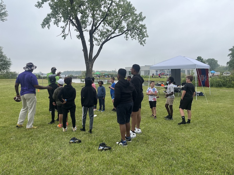 A group of children are taking direction and from two men as they wait for their next game of flag football. In the background is a tall tree in front of the shoreline of a lake.