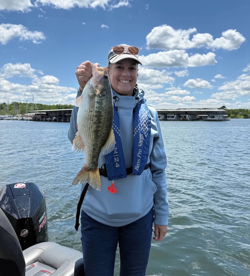 A woman standing on a boat holds up a large silver and dark green fish. In the background is a marina on a lake. The sky above is partly cloudy.