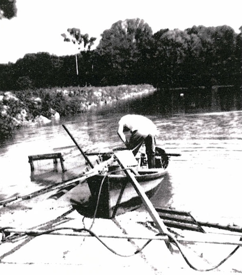 A black and white historical photo of a biologist running a boat with an electric shocking device to study fish on a lake. In the background is a shoreline against a woodland.