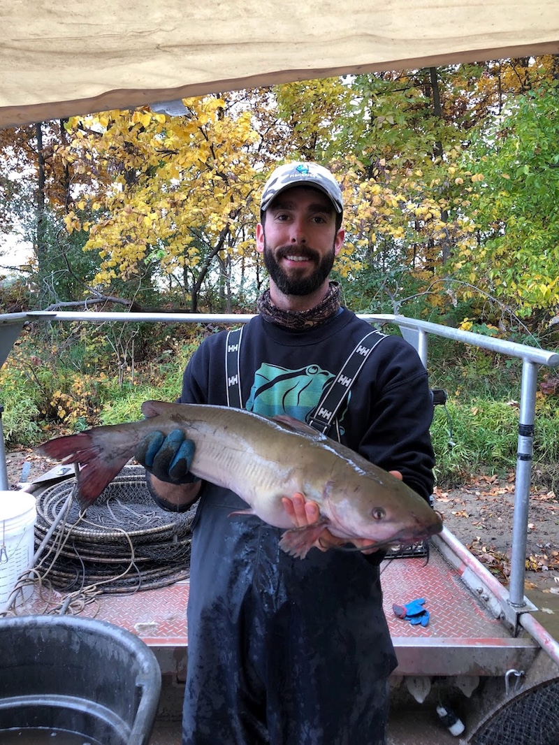 A man standing in a boat holds up a large tan, gray and silver fish with whiskers around its mouth. In the background is a shoreline with autumn trees and green vegetation.