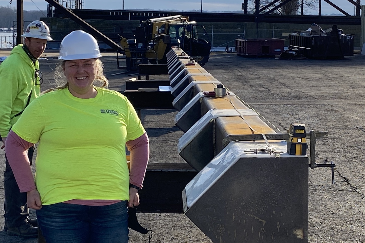 A woman wearing a hard hat stands next to a large piece of machinery. She stands at one end of a long large bar with rectangular projections at equal intervals down the length of the bar. In the background is another individual wearing a hard hat standing in a parking lot.