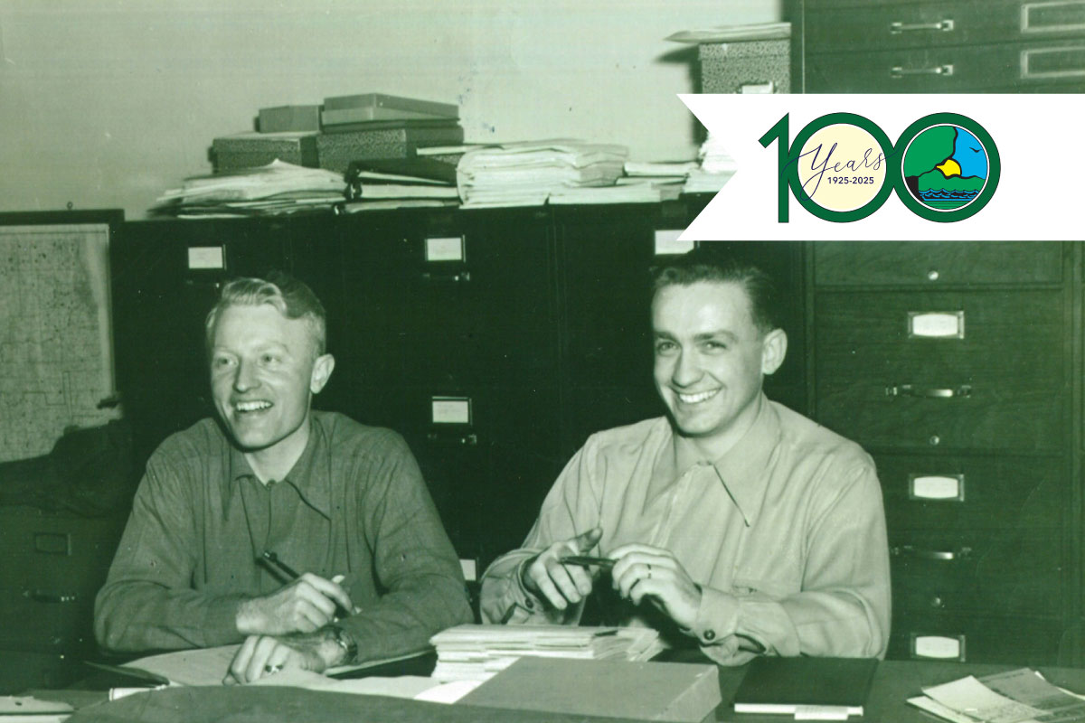 A historical black and white photo with two men sitting at a desk covered with books and writing utensils. In the background are filing cabinets. Overlaying the photo on the top right is the logo for the Illinois Department of Natural Resources 100th anniversary.