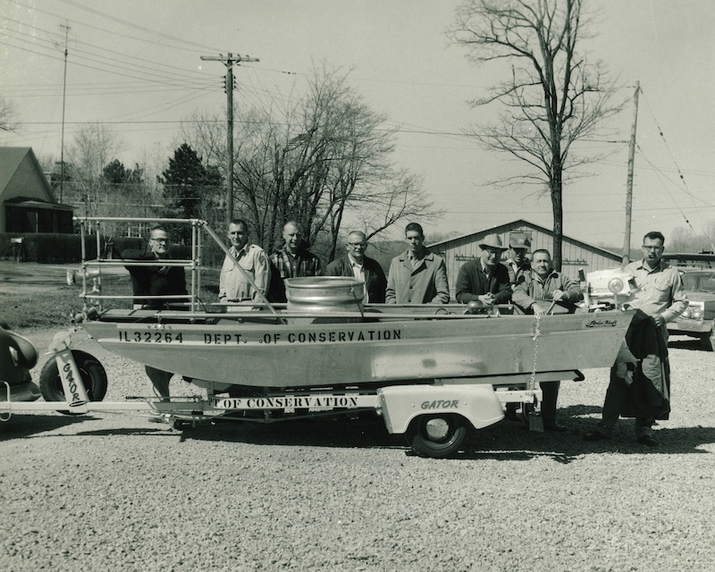 A black and white historical photo of a group of men standing behind a metal boat on a trailer in a gravel parking lot.
